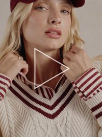 Krém színű, kötött kábelminta pulóver mély V-nyakkal és bordó csíkokkal, egy piros-fehér csíkos ing fölött viselve; bordó sapkát hord.