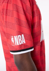 Raudonas krepšinio marškinėlių kostiumas su juodu ir baltu NBA logotipu ant rankogalių, su tamsesnio raudono atspalvio pakartojančiu „CHICAGO“ raštu.