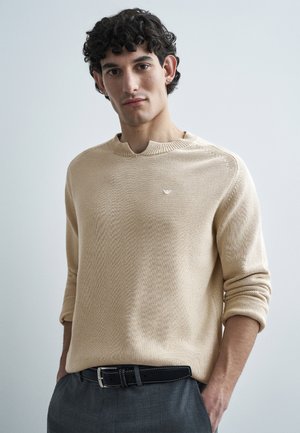 Nuori mies, jolla on kiharat mustat hiukset, beige neulepaita ja harmaat housut mustalla vyöllä, seisoo käsivarsien taskuissa.