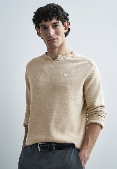 Jeune homme aux cheveux noirs bouclés portant un pull beige et un pantalon gris avec une ceinture noire, debout les mains dans les poches.