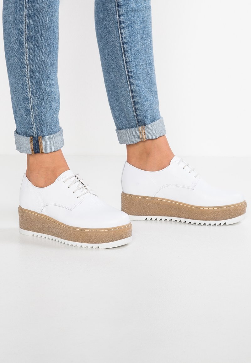 Personne portant un jean bleu clair retroussé et des chaussures à plateforme blanches à lacets, avec des semelles intermédiaires beige et des semelles extérieures blanches à crampons.