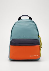 Zaino blu chiaro con tasche frontali navy e arancioni, tirazip giallo e nero, e testo bianco "S.T.H.L.M MO’P 1967" sulla tasca inferiore.