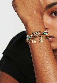Bracelet multicolore en perles avec un symbole de paix en or comme pièce maîtresse, des charms floraux et des textures variées. Porté sur un bras avec un teint de peau clair.