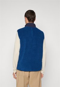 Man in een blauwe, zachte mouwloze vest over een beige langemouwen shirt en beige broek, staand met zijn rug naar de camera tegen een effen achtergrond.