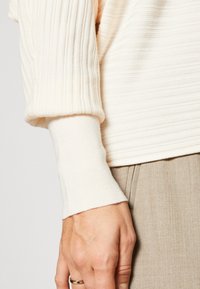 Gros plan sur une main portant une bague en or, avec une manche de pull côtelé crème et un pantalon beige visibles.