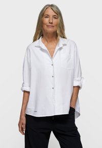 White button-up shirt made of lightweight fabric, featuring a small pocket, rolled sleeves, and curved hem, paired with black pants.