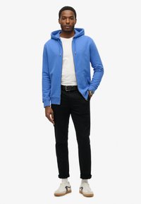 Sudadera azul con cremallera y bolsillo canguro, combinada con una camiseta blanca, pantalones negros y zapatillas blancas. El modelo está de pie frente a un fondo blanco.