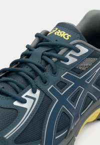 Σκούρο μπλε αθλητικό παπούτσι με διχτυωτό και συνθετικό υλικό. Διαθέτει γκρίζες λεπτομέρειες, ανακλαστικά στοιχεία και κίτρινο λογότυπο. Παραδοσιακό σχέδιο με κορδόνια.