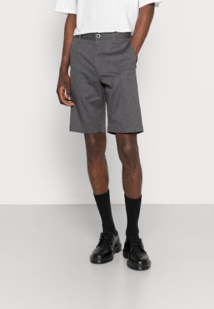 Homme portant un short gris au genou, des chaussettes noires et des chaussures noires à lacets, debout, une main dans la poche, sur un fond uni.