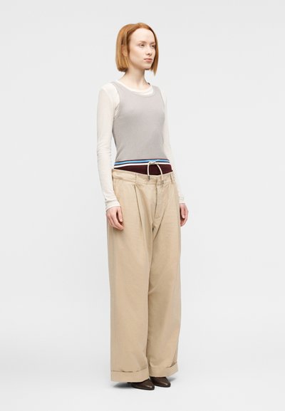 Γκρι ριγωτό αμάνικο τοπ φορεμένο πάνω από λευκή μπλούζα με μακριά μανίκια, σε συνδυασμό με μπεζ παντελόνι φαρδιάς γραμμής με ζώνη και γυρισμένα μανσέτα.