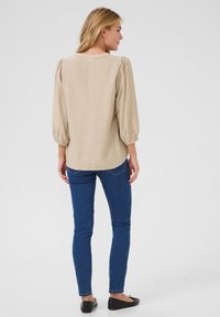 Haut beige à manches bouffantes avec un col rond, en tissu texturé, et une ligne d'ourlet légèrement incurvée à l'arrière, associé à un jean skinny bleu.