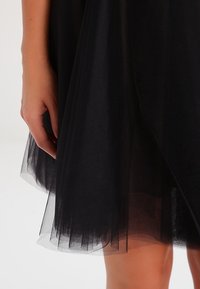 Svart tyllkjol med ett lager av genomskinligt material och fransat nederkant. Nära bild av en hand som vilar på kjolens kant, vilket visar texturen.