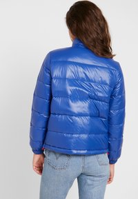 Modrá vyp Paddingová bunda s lesklým povrchem, s vodorovně prošívanými sekcemi a vysokým límcem; v kombinaci se světle modrými džíny.