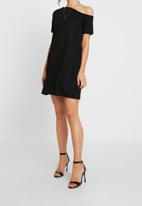 Černé šaty s odhalenými rameny z hladkého materiálu s krátkými rukávy. Vybavené černými sandály na vysokém podpatku s kotníkovými pásky.