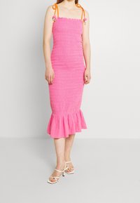 Vestido midi rosa con smocks y dobladillo con volantes, tirantes ajustables naranjas y tela texturizada. Lleva zapatos de tacón blancos con tiras.