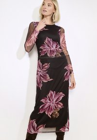 Žena v černých průsvitných dlouhých šatech s velkými růžovými květinovými vzory, která pózuje proti jednoduchému světlému pozadí.