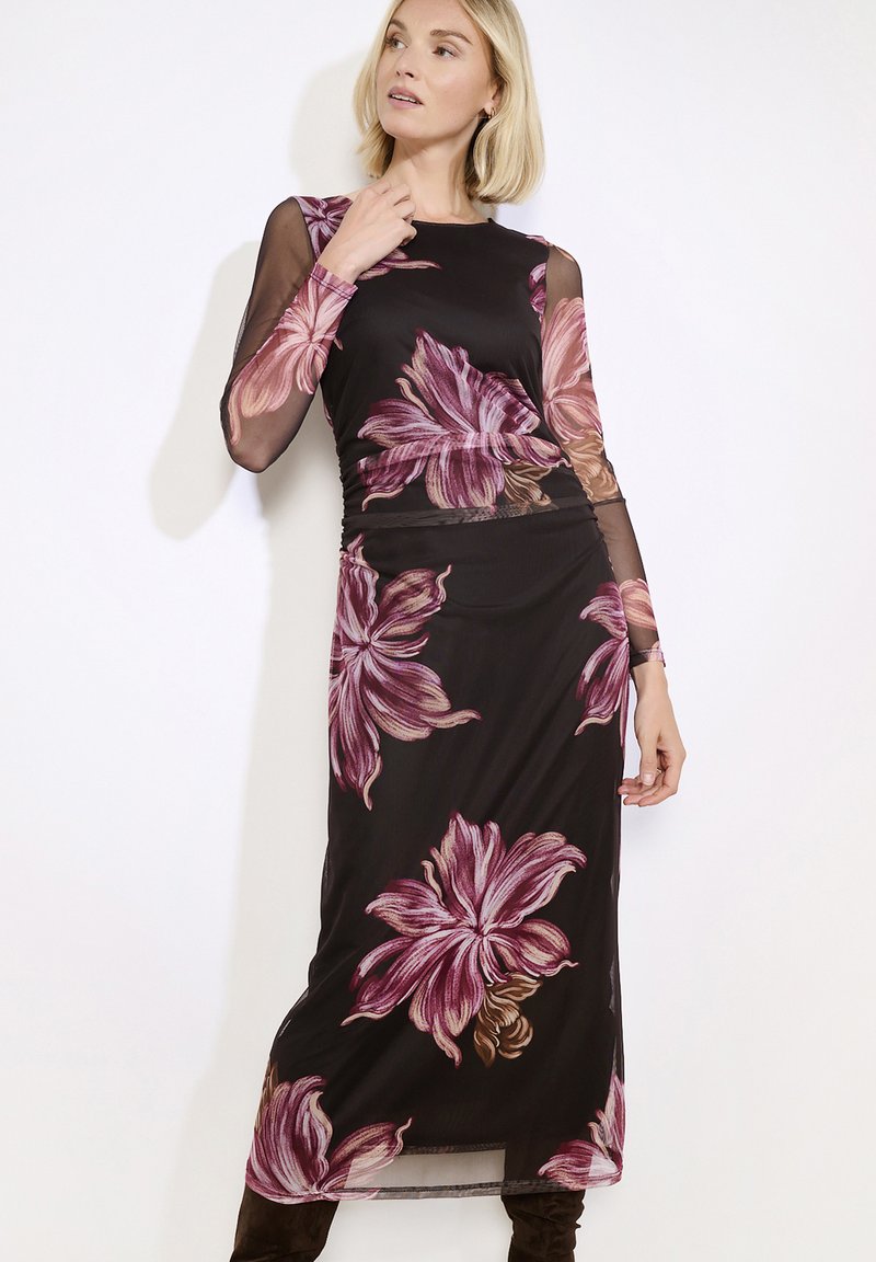 Žena v černých průsvitných dlouhých šatech s velkými růžovými květinovými vzory, která pózuje proti jednoduchému světlému pozadí.