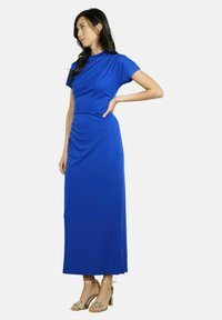 Modrá maxi šaty s krátkymi rukávmi, drapovaným výstrihom a nahrnutím na boku. Hladká tkanina s priliehavým strihom. Nosia sa s pásikavými zlatými podpätkami.