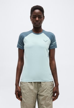 Henkilö, joka pukeutuu vaaleansiniseen ja harmaaseen lyhythihaiseen urheilupaitaan sekä beigenvärisiin housuihin, seisoo yksivärisen valkoisen taustan edessä.