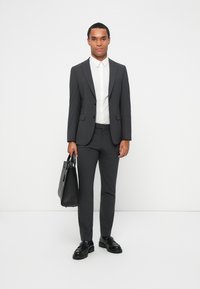 Completo grigio in tessuto texturizzato; giacca a un bottone, pantaloni slim-fit; abbinato a una camicia bianca e scarpe nere; tiene una borsa nera.