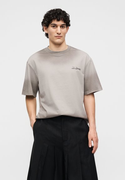 Νέος άνδρας με σγουρά μαλλιά, φορώντας ένα φαρδύ ανοιχτό γκρι T-shirt και ευρύ φαρδιά πλισαρισμένα μαύρα παντελόνια, στέκεται με τα χέρια στις τσέπες.