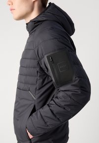 Casaco acolchoado preto com capuz, bolsos com fecho éclair e um patch de logotipo na manga. Tecido texturizado com design em canais.