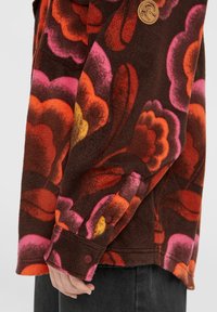 Brun fleece skjorte med store, flerfargede blomster mønstre i oransje, rosa og gul; har knapp mansjetter og en avrundet hem.