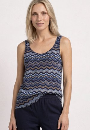 Femme aux cheveux blonds portant un haut sans manches à motif zigzag bleu, marron et blanc et un pantalon bleu marine foncé sur un fond clair uni.
