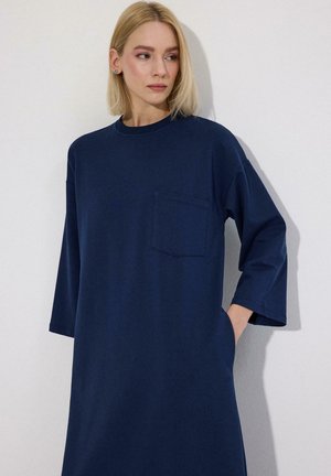 Lyshåret kvinde iført en løs, mørkeblå kjole med 3/4 ærmer og en brystlomme, stående op ad en hvid væg med den ene hånd i sin sidelomme.