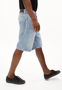 Shorts en denim bleu clair avec une coupe droite, un ourlet brut et des détails de couture visibles. Ceinture noire avec logo, associés à des baskets noires.