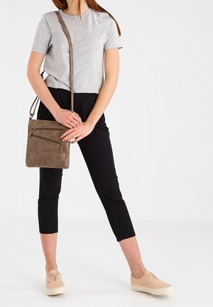 Woman wearing gray t-shirt and black cropped pants, holding a taupe crossbody bag, standing with one leg slightly bent.