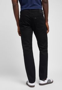 Homme portant un jean noir, une chemise bleu marine et des baskets blanches avec des bandes noires, debout de dos sur un fond uni.