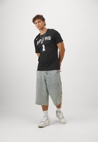 Sort T-shirt med korte ærmer med "SPURS" og nummer "1" i hvid, parret med lyseblå denimshorts og hvide sneakers.