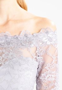 Top bez ramínek v bledě levandulovém krajkovém provedení, zdobený květinovými vzory a průsvitnými dlouhými rukávy. Jemný krajkový lem s vlnkami na výstřihu.
