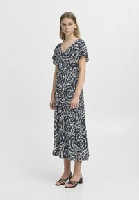 Marineblaues Kleid mit floralem weißen Muster, kurzen Schmetterlingsärmeln, V-Ausschnitt und ausgestellt geschnittenem Saum. Getragen mit schwarzen Riemchensandalen.