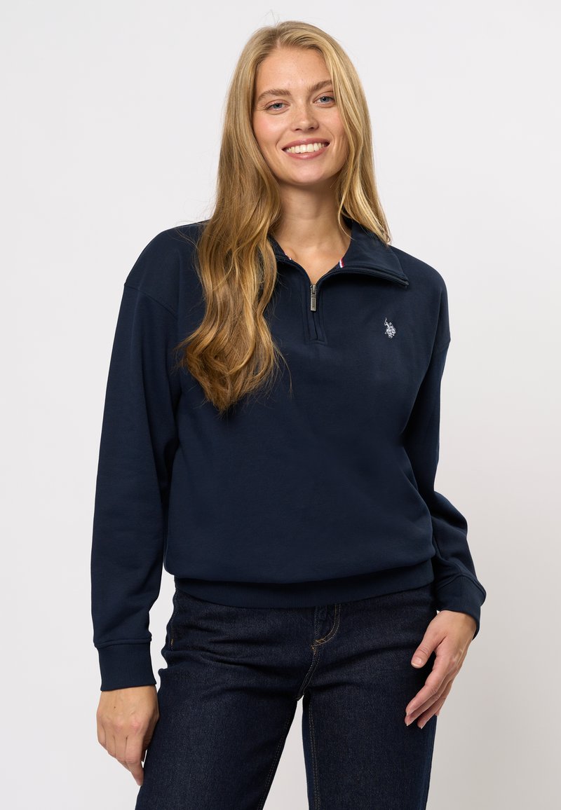 Marineblå genser med kvart-glidelås krage, ribbede mansjetter og en liten brodert logo på brystet. Laget av myk, teksturert stoff.