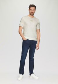 Jasny beżowy T-shirt z krótkim rękawem z prostokątnym logo, w połączeniu z ciemnoniebieskimi dżinsami i białymi sneakersami, prezentujący swobodny styl.