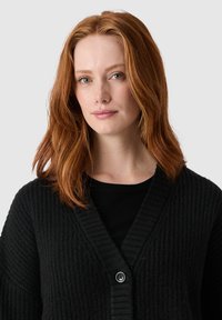 Czarny kardigan z teksturowanym wzorem dzianiny, dekoltem w serek i prążkowanymi detalami. Posiada pojedyncze zapięcie na czarny guzik.