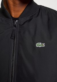 Veste bomber noire avec fermeture éclair, col côtelé et un petit logo crocodile vert et blanc sur le côté gauche. Texture de tissu lisse.