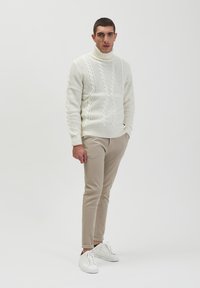 Chunky witte kabelgebreide trui met hoge kraag, gedragen met beige slim-fit broek en witte sneakers. Soh tere structuur en casual ontwerp.