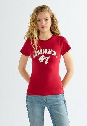 Młoda kobieta o długich blond kręconych włosach, ubrana w czerwoną koszulkę Wrangler 47 i jasnoniebieskie jeansy, stoi z rękami za plecami.