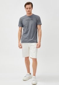 Camiseta gris de manga corta con el texto "LO SIENTO, LLEGUÉ TARDE" en negro, combinada con pantalones cortos blancos. El modelo lleva zapatillas blancas; diseño simple y casual.
