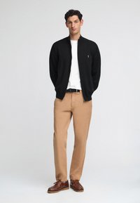 Svart zip-up jacka över en vit skjorta, tanfärgade byxor och bruna skor. Slät textur, avslappnad design, med subtil logotypdetalj på jackan.