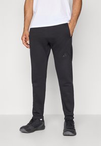 Férfi fekete Adidas melegítőnadrágban és fekete sportcipőben, az egyik kezét a zsebébe dugva áll egy sima háttér előtt.
