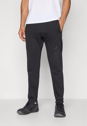 Homme portant un pantalon de survêtement noir Adidas et des baskets noires, se tenant avec une main dans la poche devant un fond uni.
