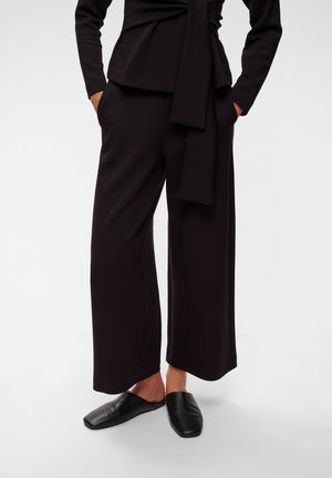 Person iført løse sorte bukser og sorte slip-on sko, stående med hænderne i lommerne mod en ensfarvet baggrund.