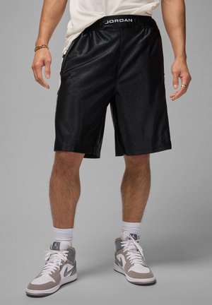 Sorte idrettsshorts laget av skinnende materiale med elastisk midjebånd som har "JORDAN"-merke. Brukt med hvite sokker og grå joggesko.