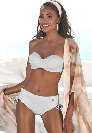 Weißes, strukturiertes Bikini-Set mit einem trägerlosen Oberteil und hochgeschnittenen Bottoms. Begleitet von einem fließenden, bunten Überwurf.