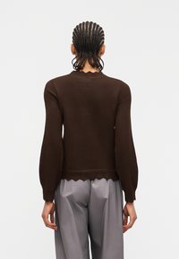 Άτομο με πλεξούδες που φοράει σκούρο καφέ πουλόβερ με μακριά μανίκια και φαρδιά γκρι παντελόνια, γυρισμένο με την πλάτη προς το μέρος ενός απλού φωτεινού φόντου.