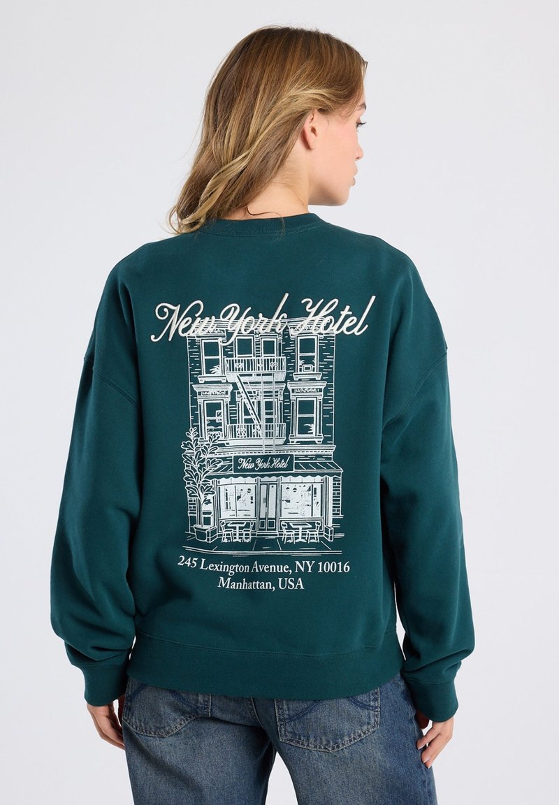 Sudadera color teal con un gráfico blanco de un edificio de hotel en la parte posterior, con "Hotel Nueva York" y detalles de la dirección debajo. Material suave, ajuste relajado.